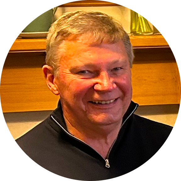 Frank Adam, a smiling older man with short, light-colored hair, wearing a black zip-up sweater indoors.